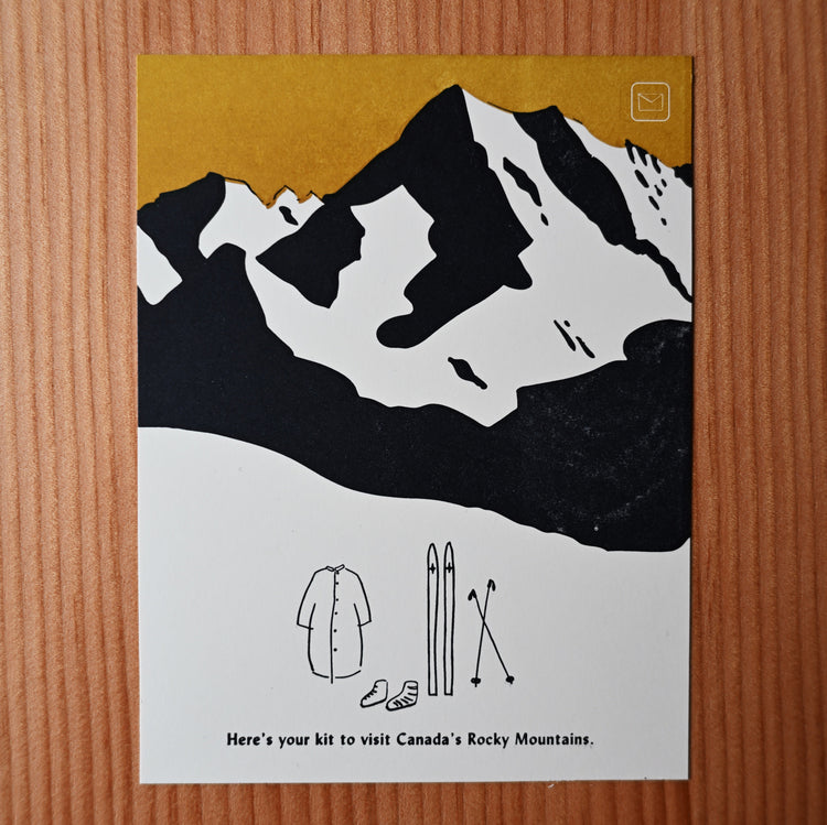 A postcard showing a striking view of the Canadian Rocky Mountains with a small hand-drawn graphic beneath it of some skis, poles, boots and a winter parka. The art print is in 2 colours, ochre and a warm navy.