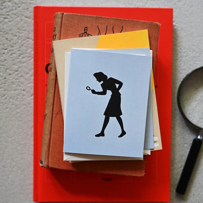 A stack of vintage books and papers on a textured light gray surface, featuring a light blue letterpress greeting card with a black silhouette of the girl detective Nancy Drew holding a magnifying glass, styled with a red book and a magnifying glass prop.